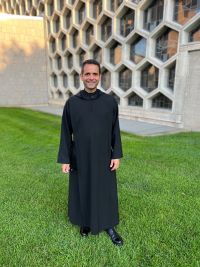 Fr in black religious robes standing outside in the grass smiling