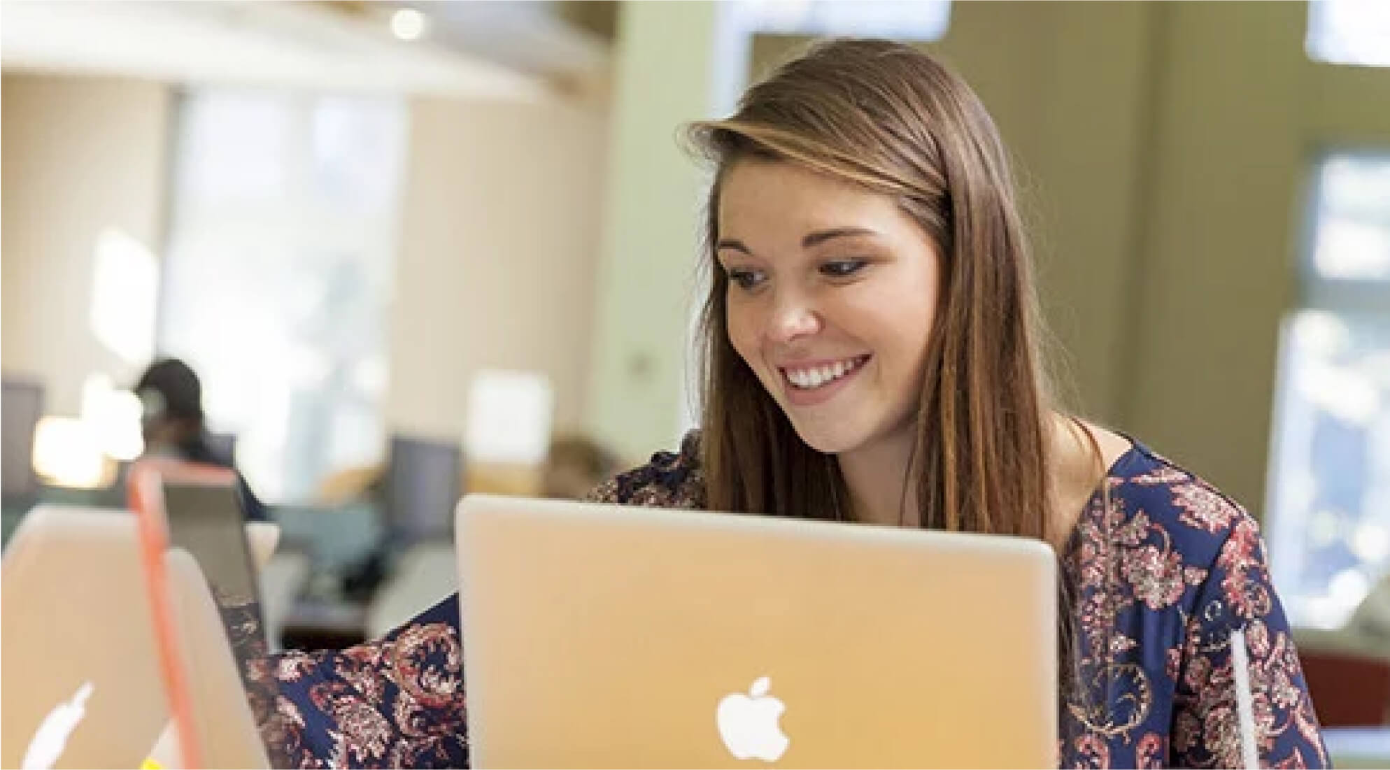 A woman with long brown hair, wearing a floral blouse, is smiling while using a silver MacBook laptop. She is seated in a well-lit room with large windows in the background, and additional laptops and people can be seen in the blurred background.