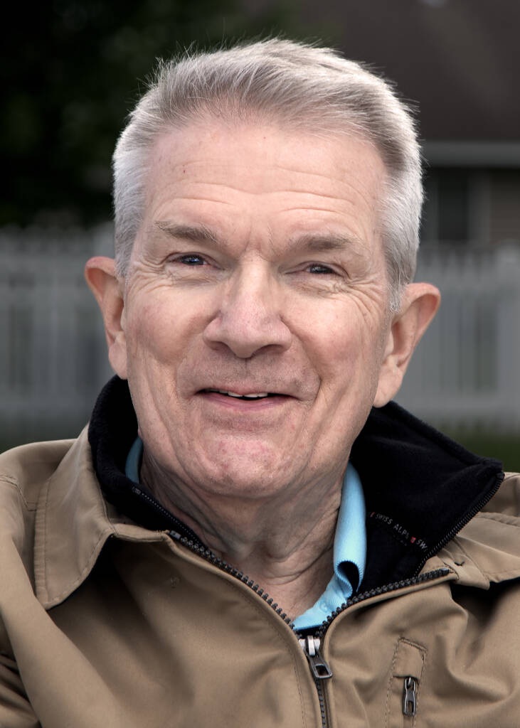 An older man with short gray hair smiles softly at the camera. He is wearing a light brown jacket over a blue shirt, and the background shows a white fence and greenery.