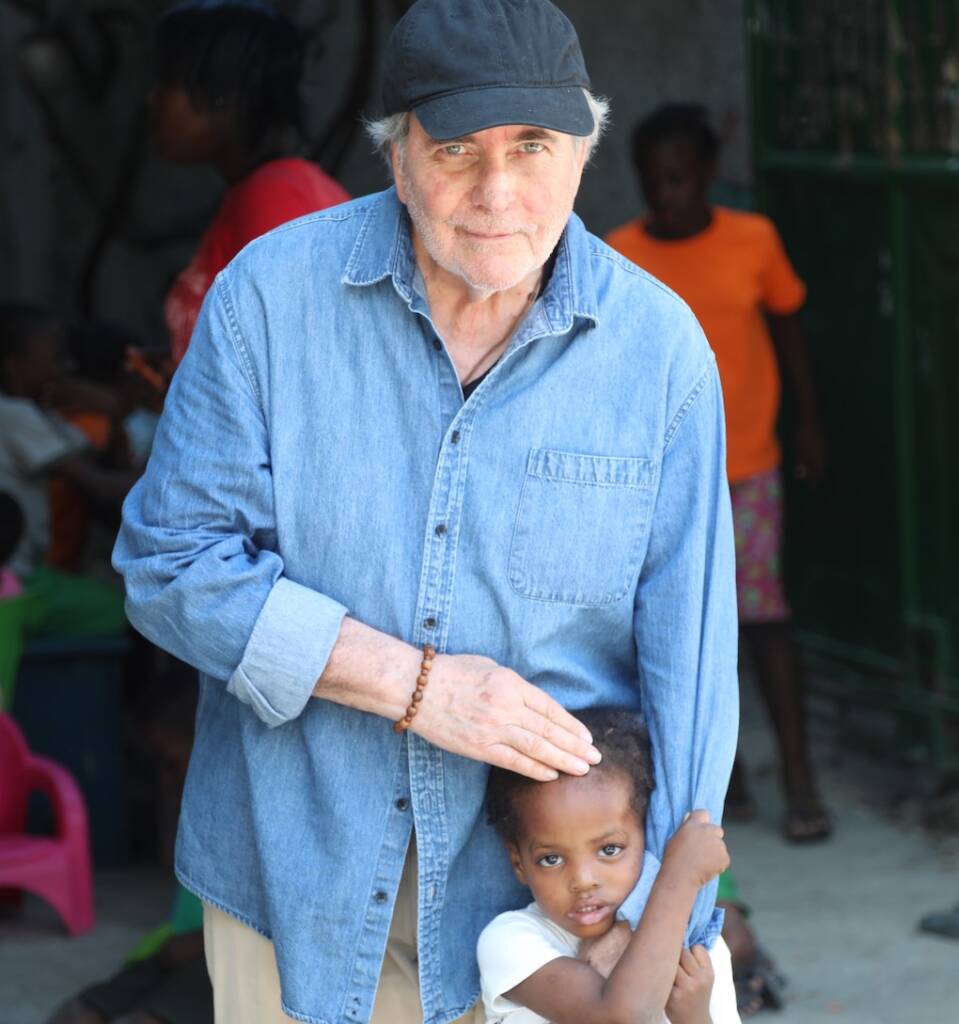 An older man in a denim shirt and cap gently places his hand on a small child who clings to his leg. The child looks at the camera, while other people are visible in the background.