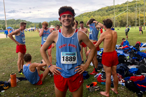 A smiling male cross country runner stands with his hands on his hips, wearing a red headband and a blue "Saint John's" singlet with the number 1889. Behind him, other runners in similar attire are stretching, talking, or resting on the grass near trees and equipment.