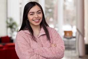 A young woman with long dark hair smiles confidently at the camera with her arms crossed. She is wearing a light pink, fuzzy sweater. The background is softly blurred, showing an indoor setting with large windows and some furniture.