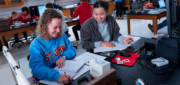 Two students sitting at a lab table, smiling and working on a computer with notebooks and scientific equipment in front of them. One wears a St. John's sweatshirt, and the other a flannel shirt. Other students are visible in the background working at separate tables.