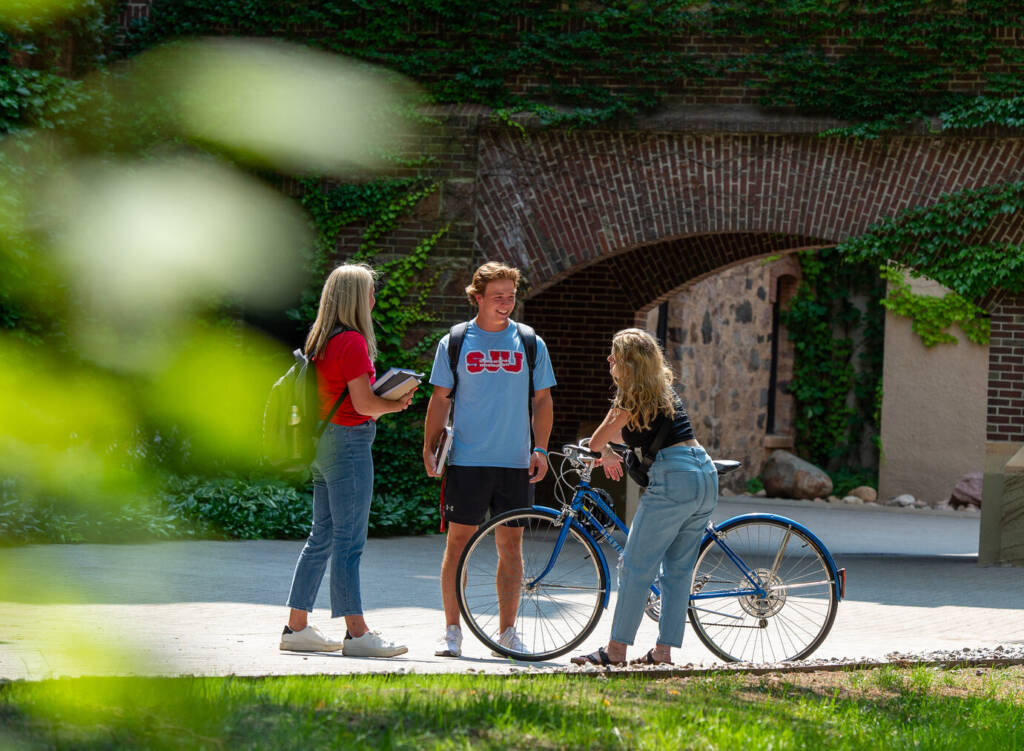 Three students have a conversation outdoors on a college campus. One holds a backpack, another carries a notebook, and the third stands with a bicycle. They are near a brick archway covered in ivy and leafy plants can be seen in the foreground.