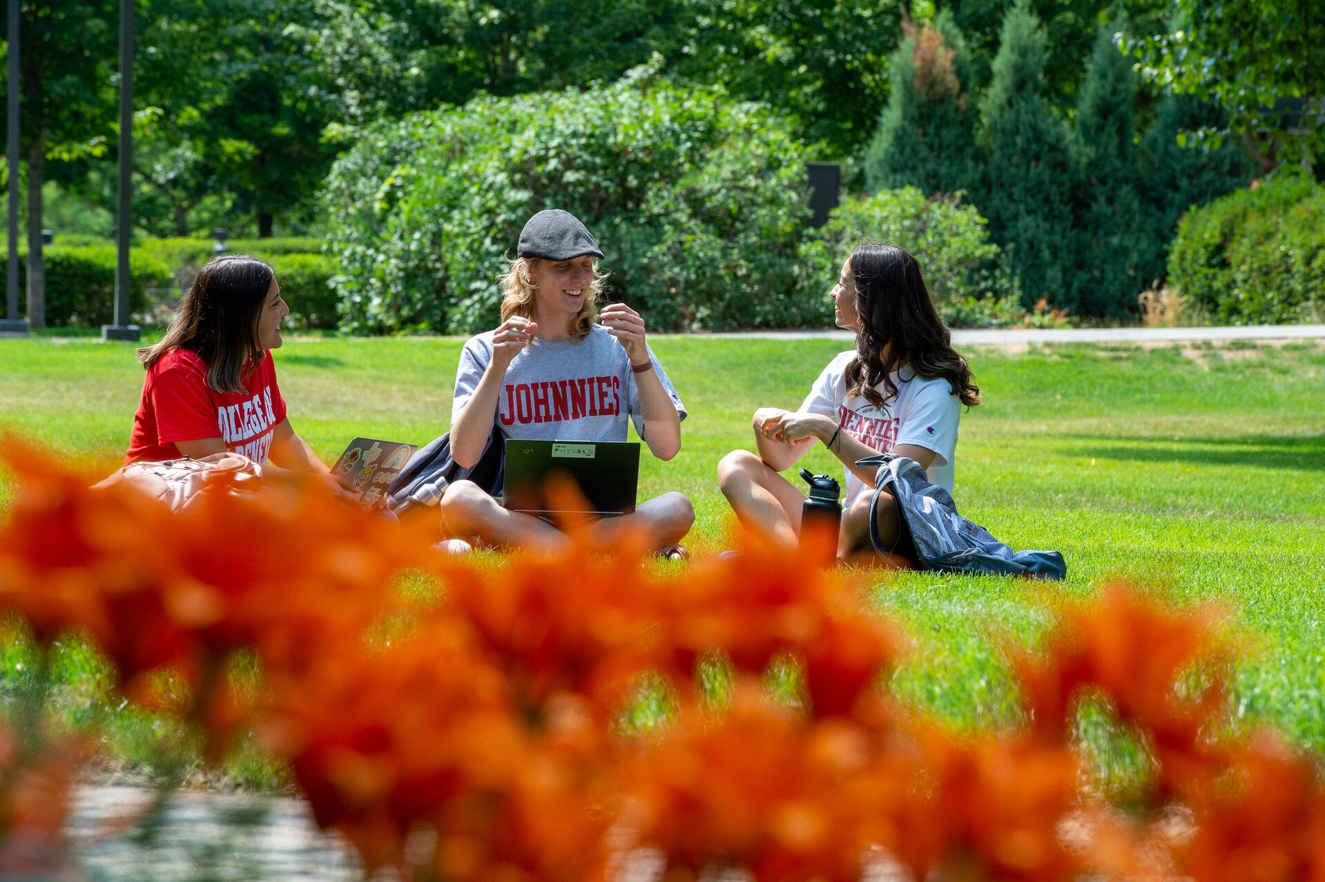 Three college students sitting on a grassy lawn, surrounded by greenery, wearing "Johnnies" and "Albany" shirts, enjoying an outdoor conversation; one with a laptop, another holding a book, and orange flowers in the foreground.