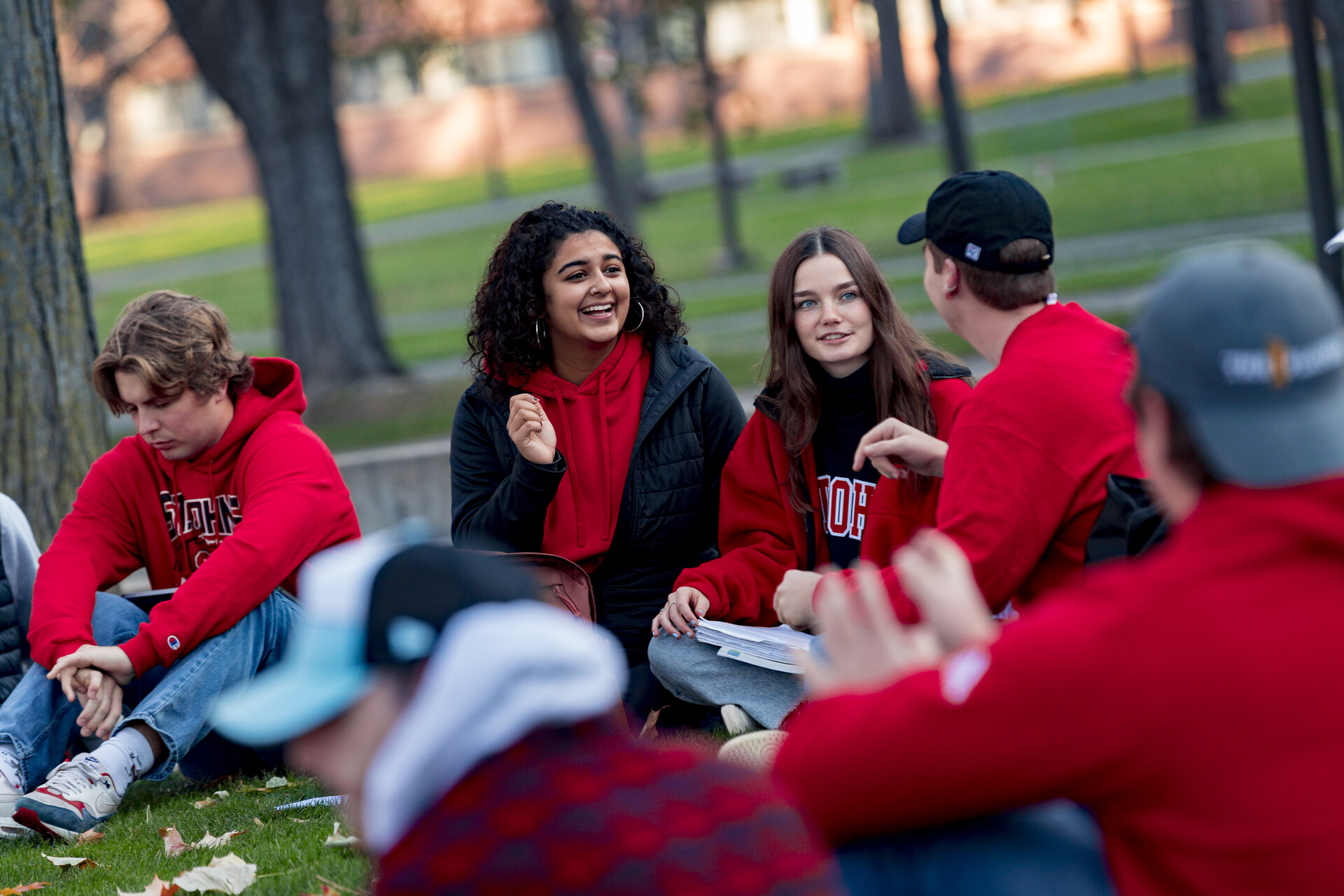 A group of students, wearing casual clothing and some in red hoodies, sit in a circle on the grass outside, engaging in conversation and smiling. Trees and a brick building are visible in the background. The scene suggests a relaxed and cheerful gathering.