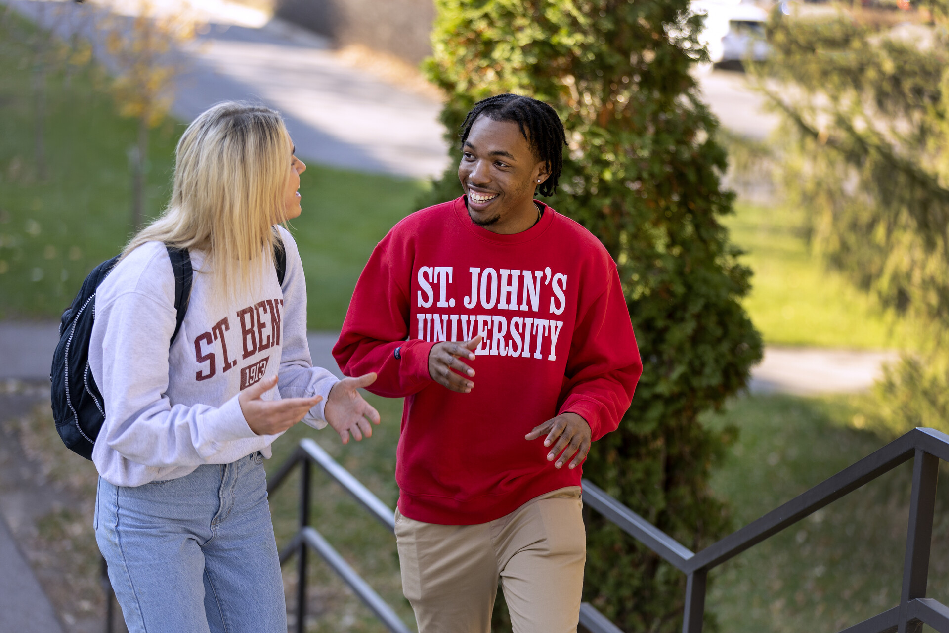 Two students, one wearing a "St. Ben's" sweatshirt and the other a "St. John's University" sweatshirt, are smiling and talking while walking up outdoor steps on a college campus. The setting includes trees and pathways, suggesting a pleasant fall day.