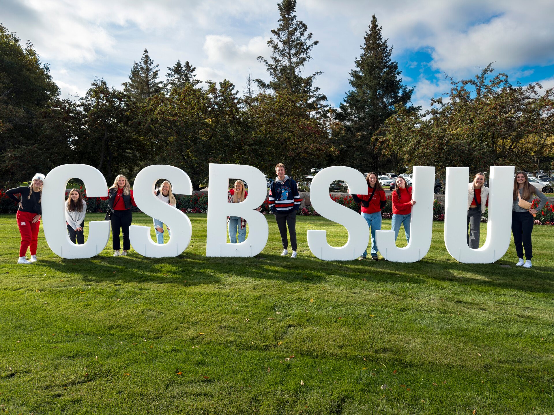 A group of people is standing on a grassy field, each person leaning against or standing beside large white letters that spell out "CSBSJU." Trees and a partly cloudy sky are in the background. The group appears to be enjoying a sunny day outdoors.