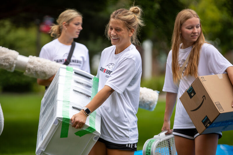 Three young women are outside carrying boxes and items, including a fan and rolled-up carpet. They are casually dressed in matching white shirts and shorts, engaged in what looks like moving or organizing activities on a grassy area.