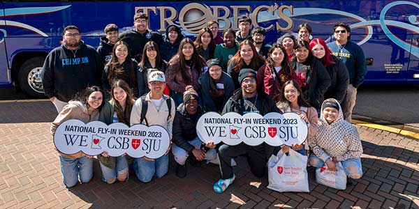 A diverse group of smiling people poses in front of a blue bus. Some hold signs reading "National Fly-In Weekend 2023" with heart symbols and university initials. Bright, sunny day on a brick-paved surface.