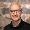 A person with glasses and a shaved head is smiling at the camera. They are wearing a black collared shirt, and the background is a textured stone wall.