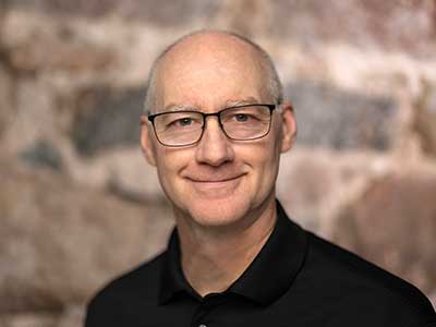 A man with glasses and a bald head smiles softly against a blurred stone background. He is wearing a black collared shirt.