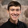 A person with short dark hair, smiling, is wearing a black shirt. The background features a blurred stone wall.