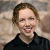 Woman with curly hair smiling, wearing a black shirt. She is in front of a blurred, neutral-toned background.