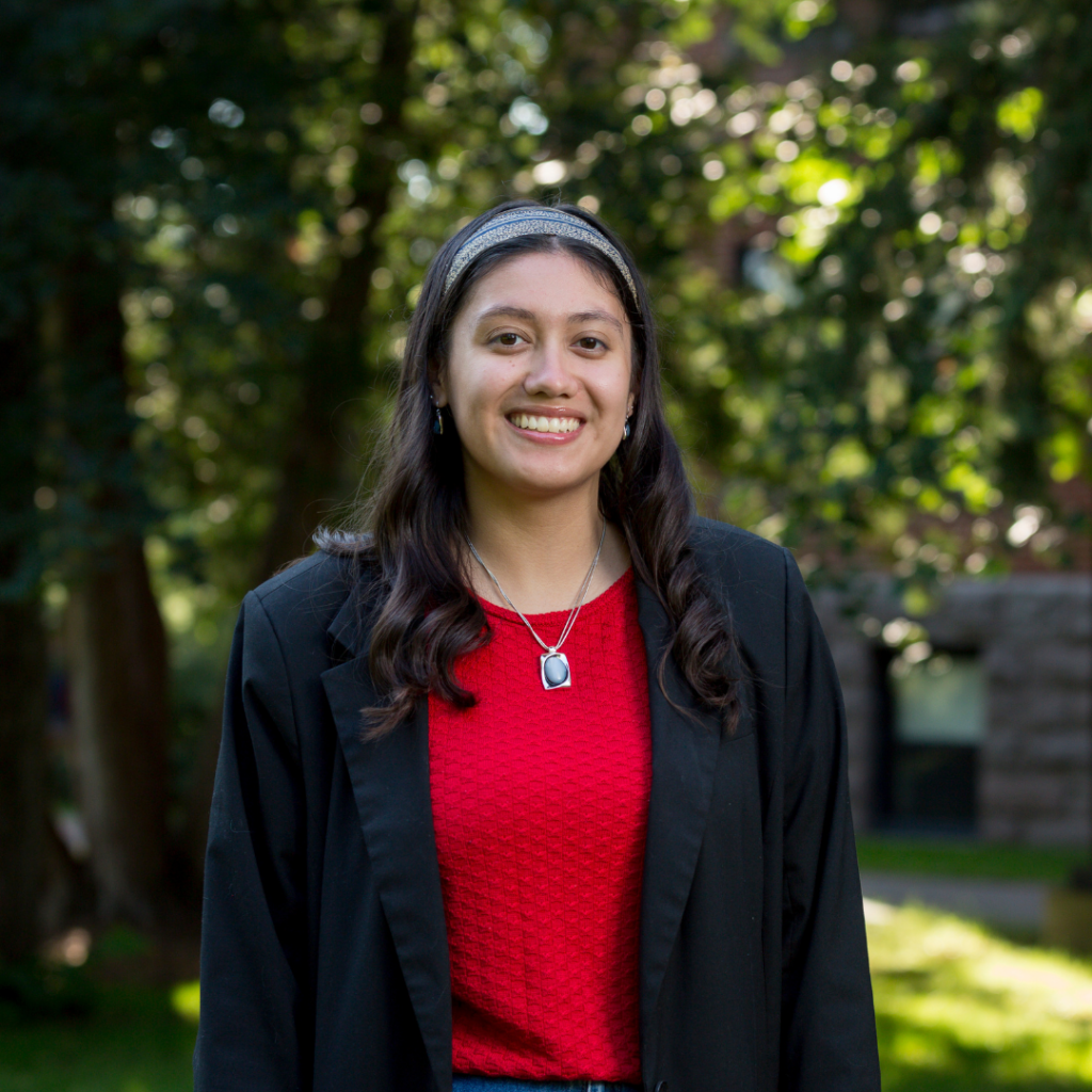 A young woman smiles at the camera, standing outdoors with trees and sunlight in the background. She is wearing a red textured shirt, a black blazer, a headband, and a necklace with a rectangular pendant.