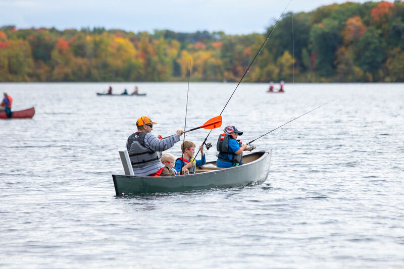 A family of four in life jackets is fishing from a small boat on a lake. A forest with autumn-colored trees lines the shore in the background. Other boats and canoes are visible on the water.