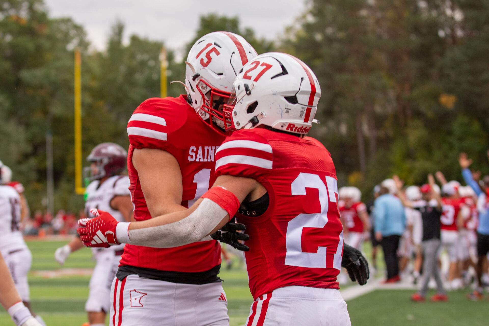 Two football players in red jerseys with white helmets embrace on a field during a game. Their jerseys display numbers 15 and 27. Other players and spectators are visible in the background amidst greenery.