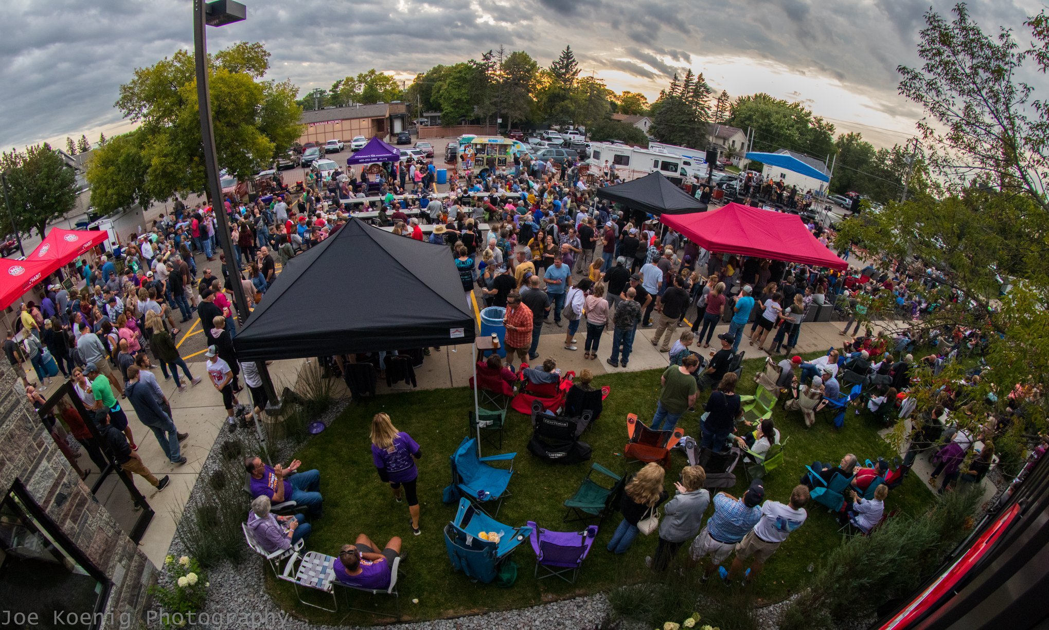 A large outdoor gathering with people standing and sitting under various colored canopies with tables. It's a lively scene, suggesting a community event or party, surrounded by trees and a few buildings. Cloudy sky overhead.