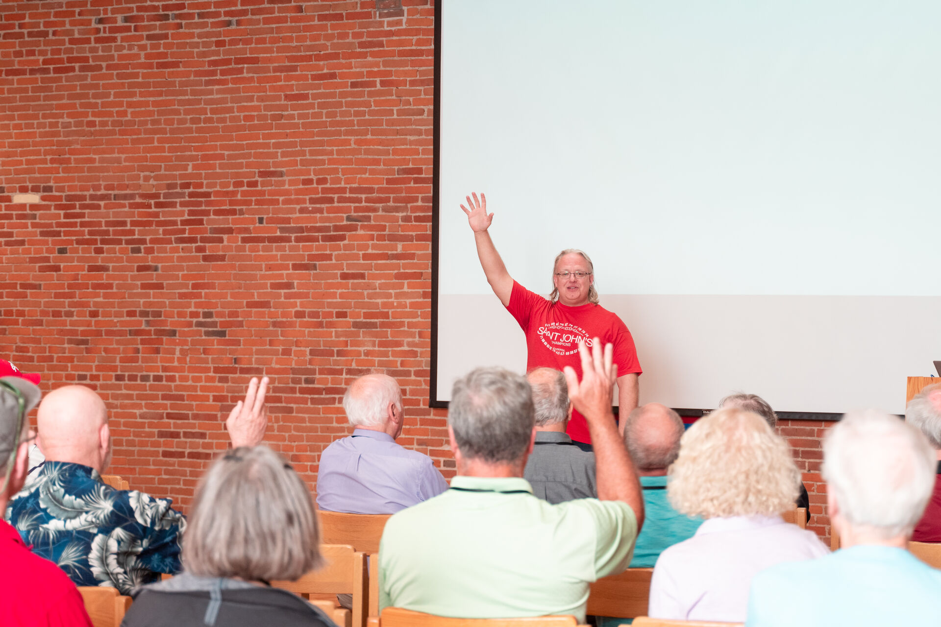 A person in a red shirt raises their hand while speaking to an audience seated in front of them. The audience, comprised of people with various clothing, sit in folding chairs. A brick wall and a projection screen are visible in the background.