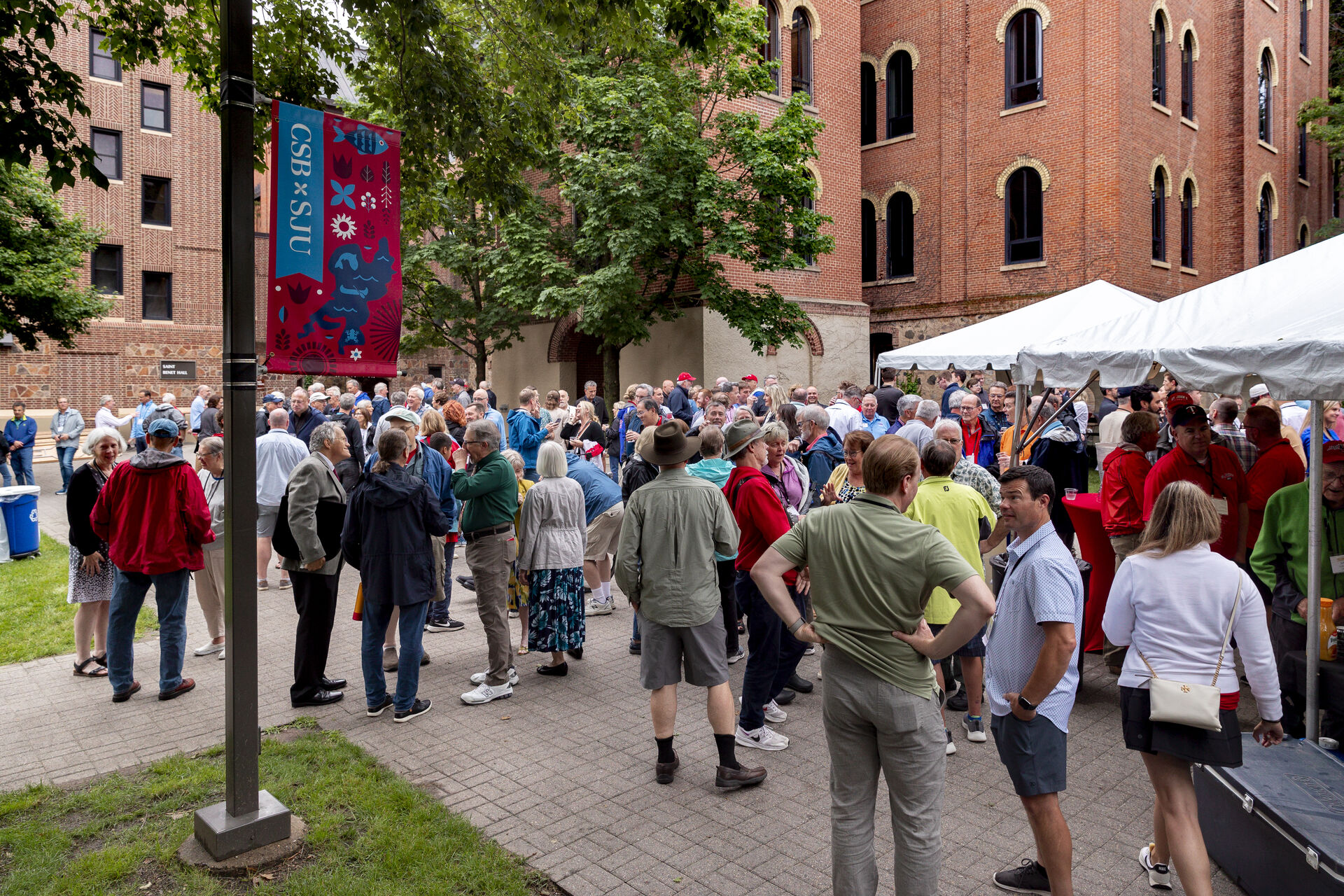 A large group of people gathered outside near a building with red brick walls, standing and interacting under trees and a white tent. A colorful event banner is on a lamp post, and attendees are dressed casually in various colors.