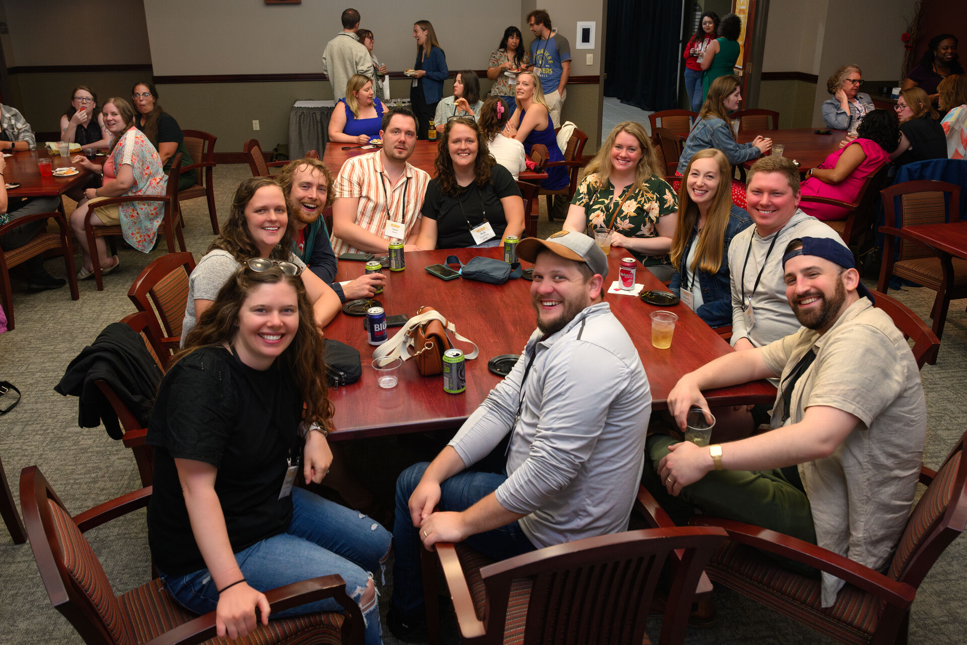 A group of people smiling and sitting around a wooden table at an indoor gathering. Some hold drinks, and they appear to be enjoying a casual social event. Other groups and tables are visible in the background.