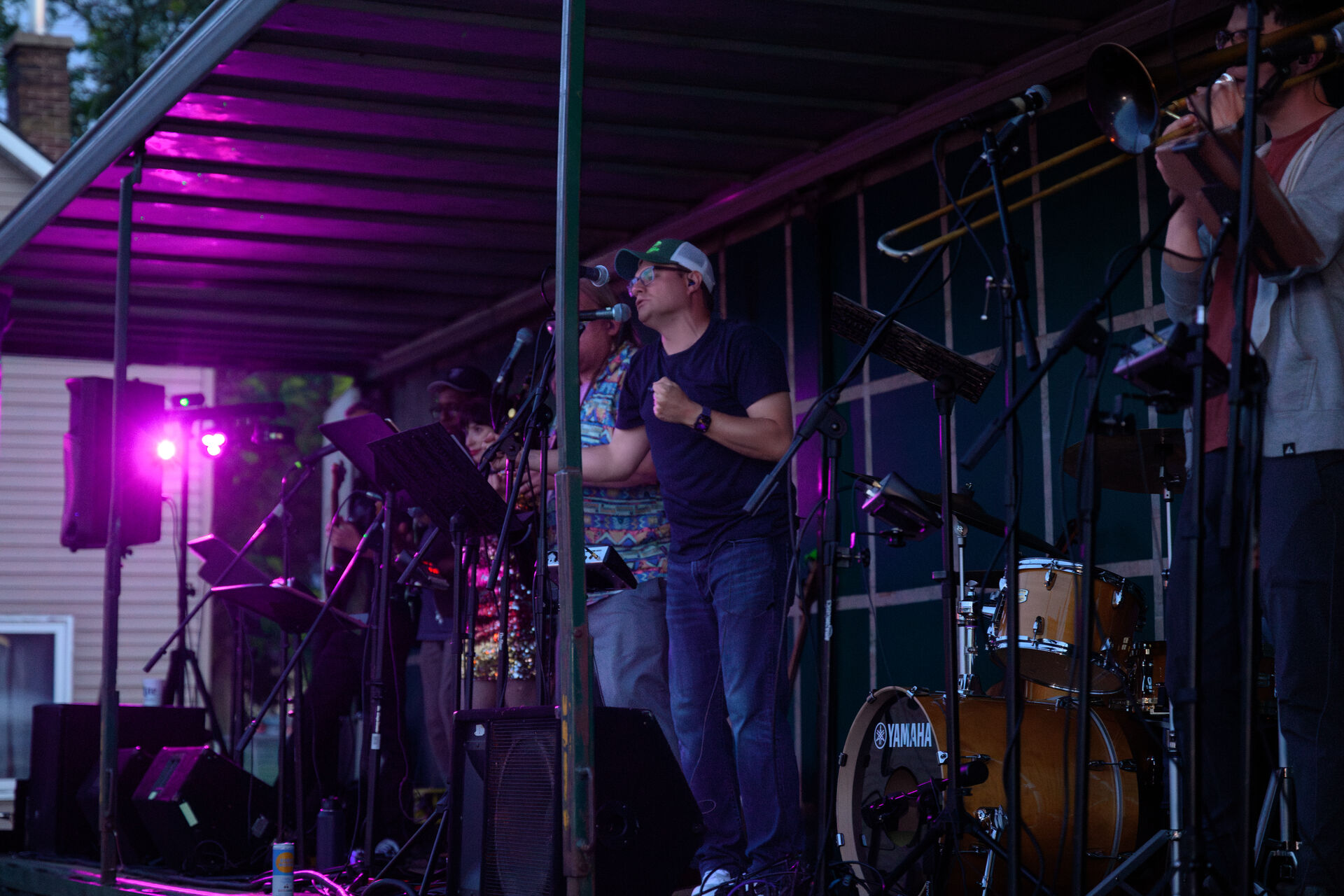 A band performs on an outdoor stage, with various musicians playing instruments like drums and a trombone. The scene is lit by magenta stage lights, and a singer at the center is holding a microphone, with buildings visible in the background.