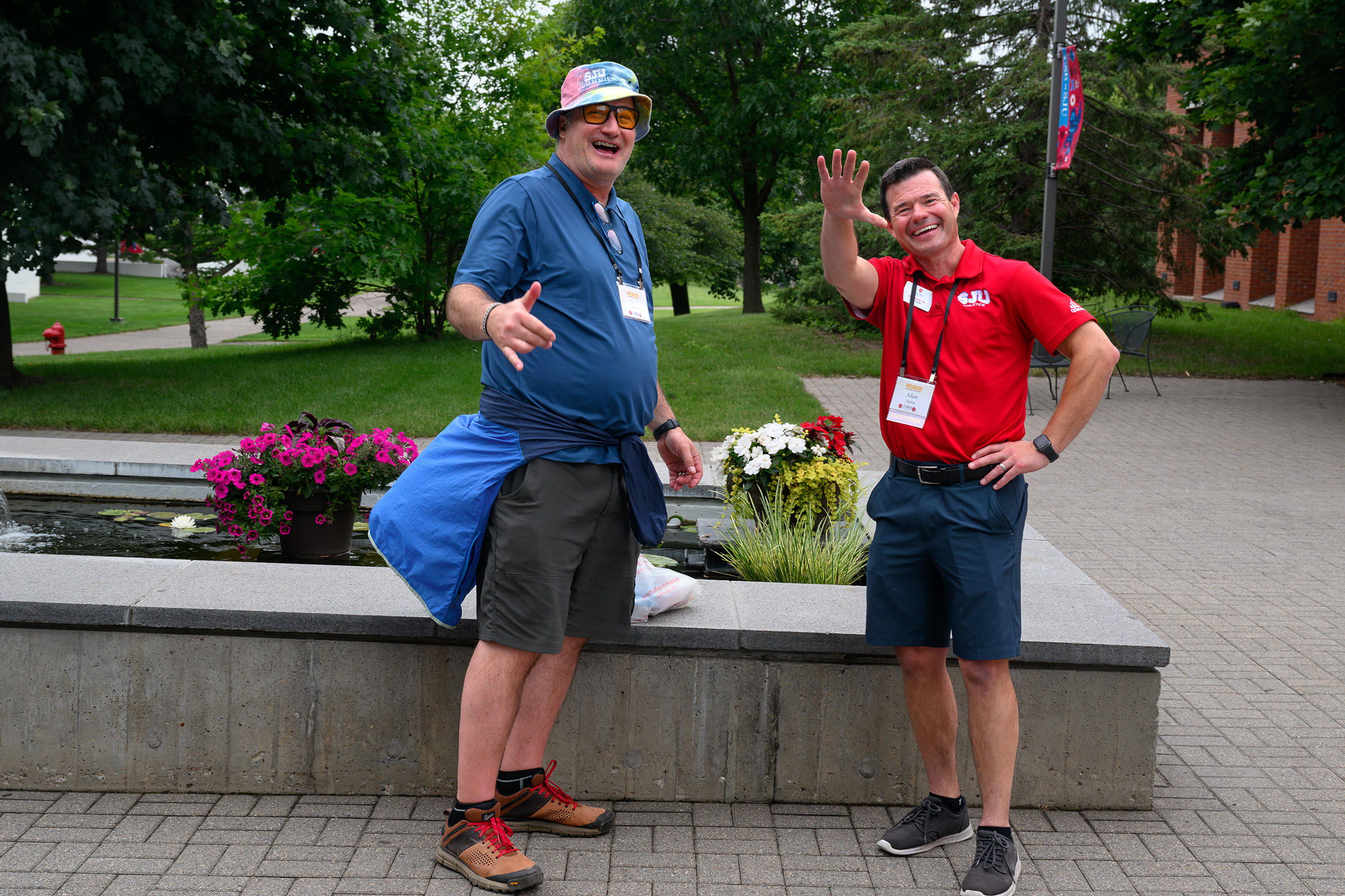 Two men are posing outdoors near a concrete planter with flowers. One wears a colorful hat and sunglasses, with a blue jacket tied around his waist. The other is in a red polo shirt with a lanyard. Both are smiling and waving. Trees are in the background.