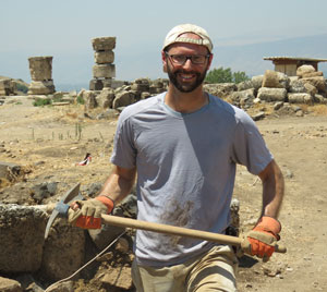 A person in a light gray shirt and cap, smiling while holding a pickaxe, stands at an archaeological site with ancient stone ruins in the background. They are wearing gloves and appear to be working outdoors under a clear sky.