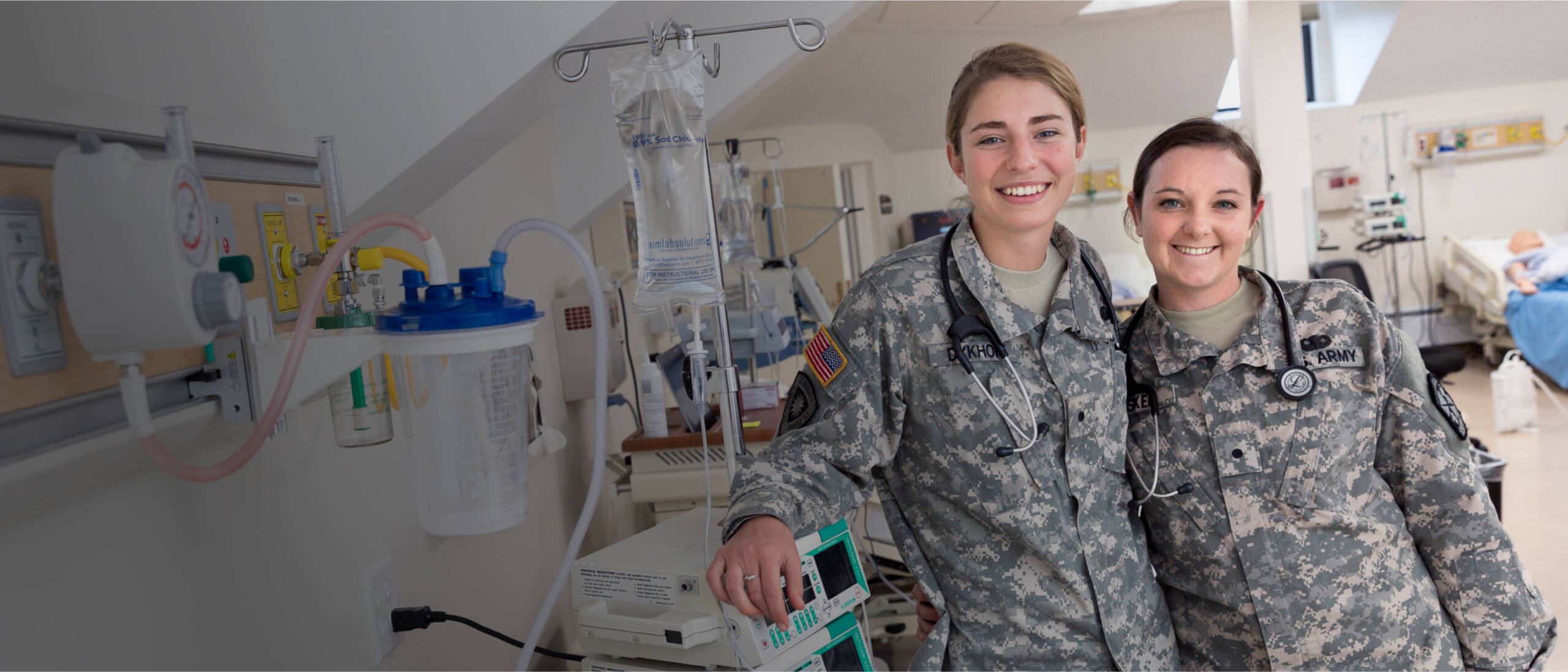 Two smiling uniformed military healthcare professionals stand side by side in a medical facility. Both are wearing camouflage uniforms with stethoscopes around their necks. Medical equipment and a patient bed are visible in the background.