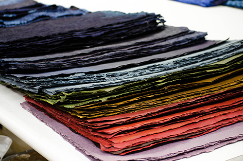 A close-up of neatly stacked, textured sheets of handmade paper in various colors, including shades of red, orange, green, blue, and purple, displayed on a white surface. The papers appear to be rough-edged, showcasing their artisanal quality.