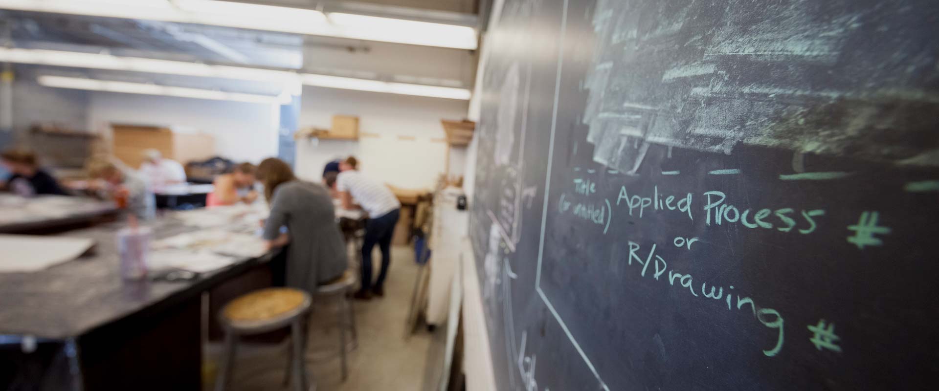 A classroom with students engaged in various activities around tables is seen in the background. In the foreground, a chalkboard displays the words "Applied Process or R/Drawing." The room appears to be well-lit with fluorescent lighting.