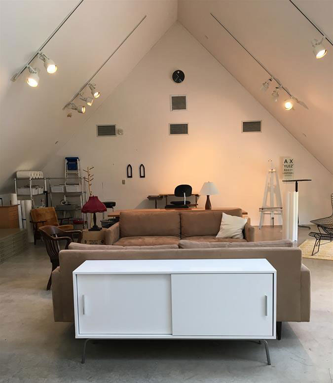 A spacious modern living room with a high ceiling and triangular shape. It features a brown sofa, white cabinet, and various chairs. Shelves and a desk are in the background, with track lighting and a minimalist design.