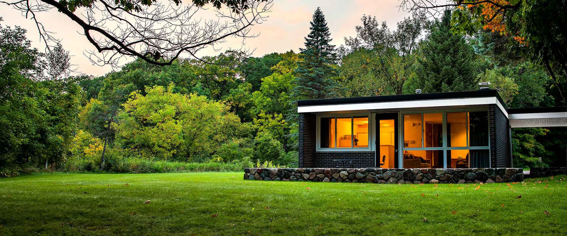 A modern, single-story house with large windows sits in a grassy clearing surrounded by lush trees. The warm glow from the windows contrasts with the green and autumn-colored foliage under a dusk sky.