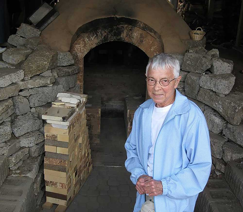 An elderly person wearing glasses and a light blue jacket stands in front of a brick kiln. The kiln is surrounded by various stones, and a basket is visible in the background.