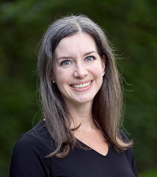 A smiling woman with shoulder-length brown hair mixed with some gray stands in front of a blurred green background. She is wearing a black V-neck top and pearl earrings.