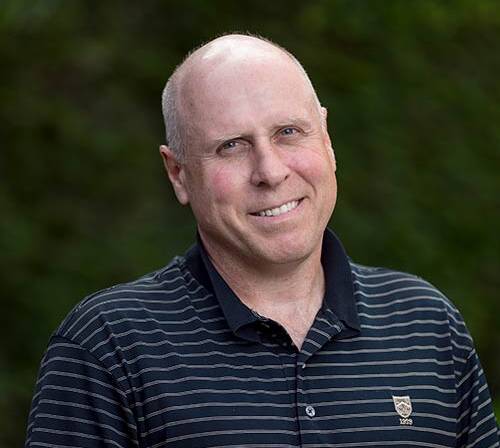 A middle-aged man with a bald head smiles at the camera. He is wearing a black, short-sleeved, striped polo shirt with a small emblem on the left side. The background is blurred greenery.