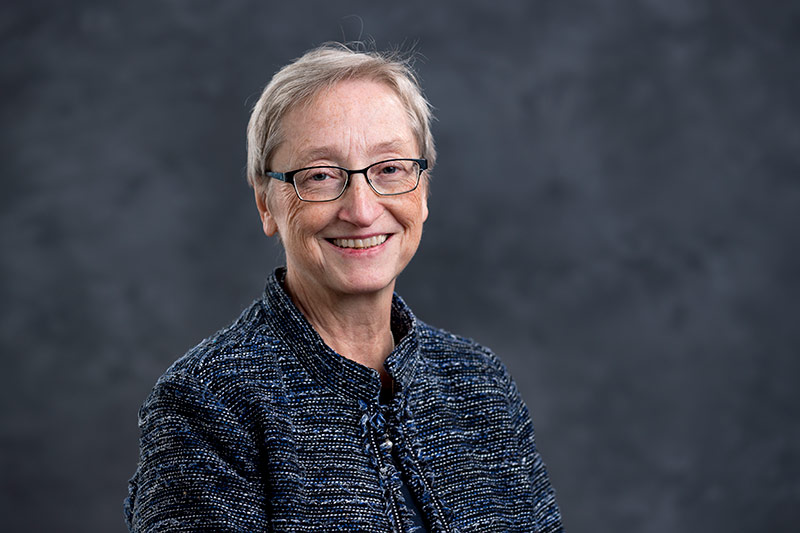 A smiling elderly person with short grey hair and glasses is seen in a professional portrait. They are wearing a dark, textured jacket and standing against a plain, dark grey background.