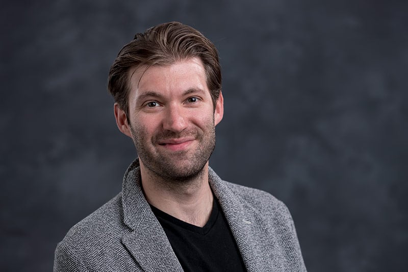 A man with short brown hair and a beard is smiling at the camera. He is wearing a black T-shirt under a gray blazer. The background is a dark, blurred texture.