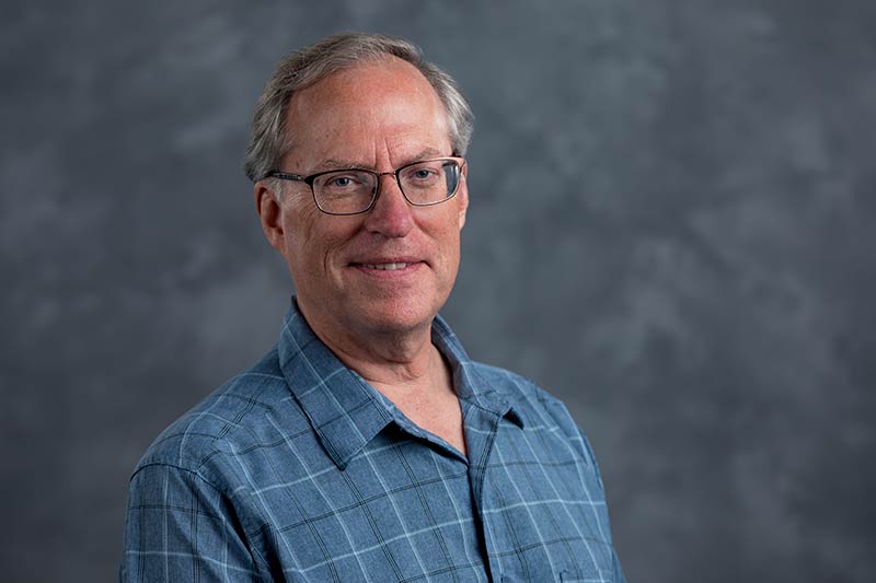 A middle-aged man with glasses and short, light-colored hair is smiling softly. He is wearing a blue plaid shirt and standing against a blurred gray background.