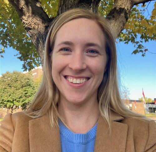 A smiling woman with long blonde hair wearing a tan coat and a blue sweater stands in front of a tree with green and yellow leaves. The background includes a building and a clear blue sky.