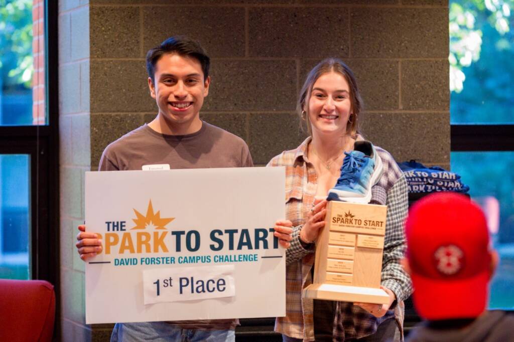 Two people smiling and holding a "1st Place" sign and a wooden trophy with a blue shoe on it. The sign reads "The Park to Start David Forster Campus Challenge." They are indoors near a brick wall and large window.