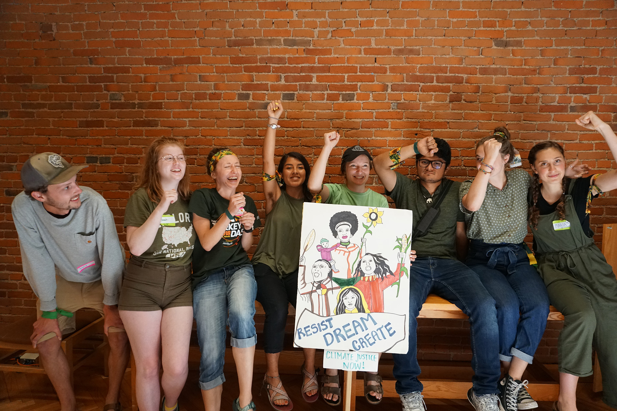 A group of eight people, casually dressed, sit and stand together in front of a brick wall, raising their fists in a jubilant manner. They hold a colorful sign that reads "Resist Dream Create" and "Climate Justice Now!" with illustrations of activists.