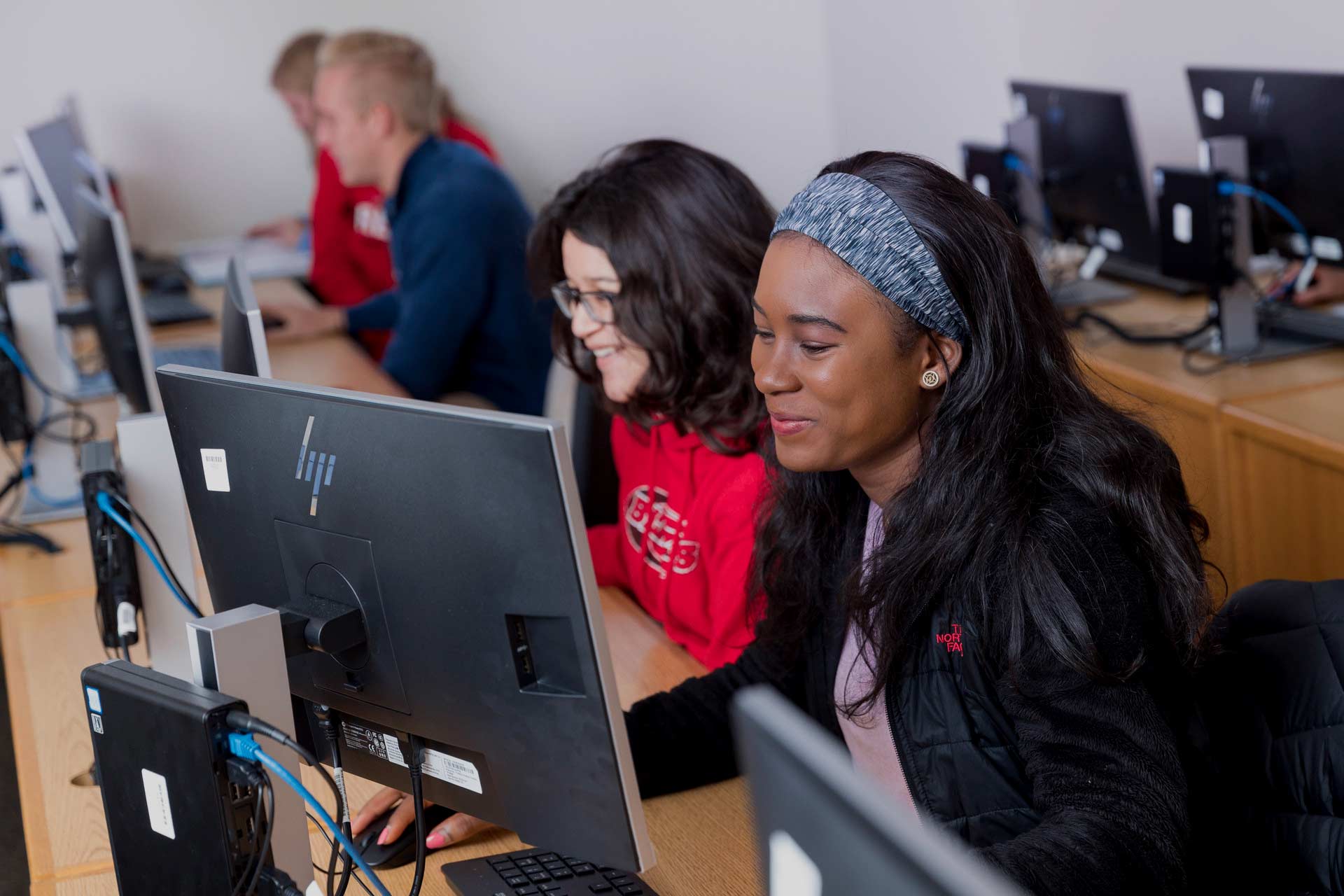 A group of students sits at a row of computers in a classroom. Two women in the foreground collaborate, one in a red sweatshirt and glasses, and the other in a black jacket and headband. More students work at computers in the background.