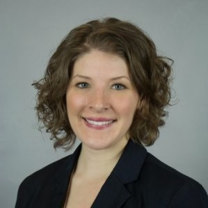 A woman with shoulder-length curly brown hair, light skin, and blue eyes smiles at the camera. She is wearing a black blazer and a light-colored top. The background is plain and gray.