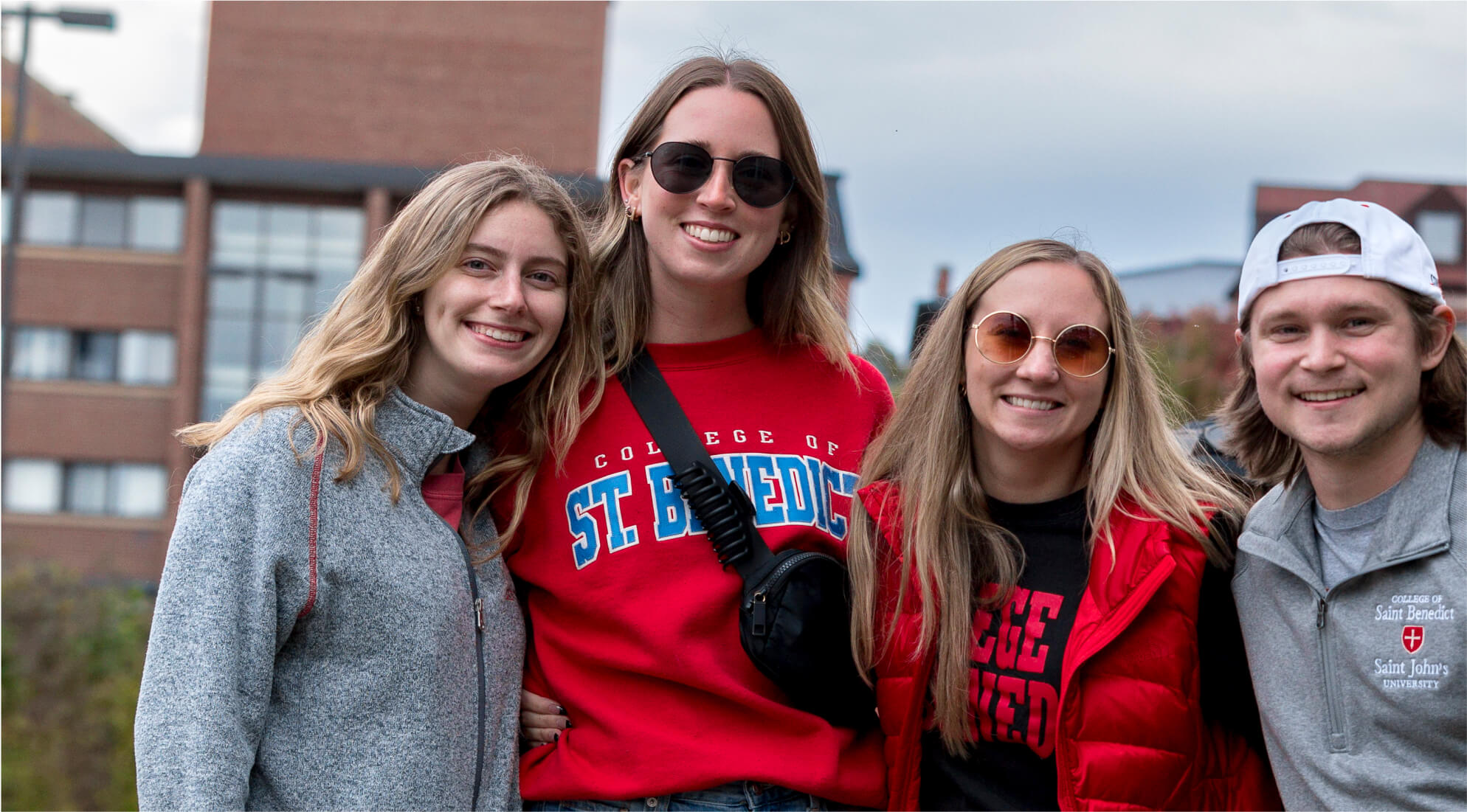 Four smiling young adults pose together outdoors, with buildings in the background. Three of them are wearing "College of St. Benedict" gear, including sweatshirts and jackets. The weather appears cool, as they are dressed warmly.