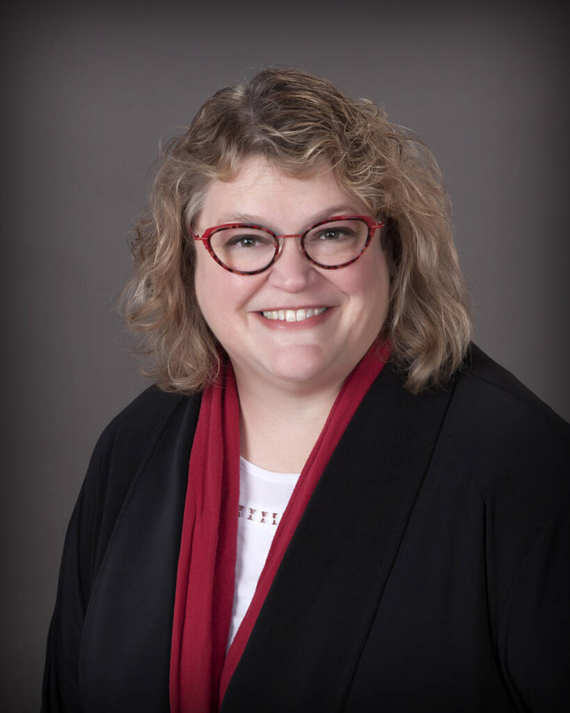 A person with shoulder-length curly hair smiles while wearing glasses and a black outfit with a red scarf. The background is a plain gray gradient.