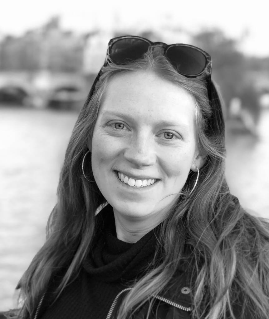 Black and white photo of a smiling woman with long hair, hoop earrings, and sunglasses resting on her head. She is wearing a black top with a high collar, standing outdoors with a blurred background showing a river and bridge.