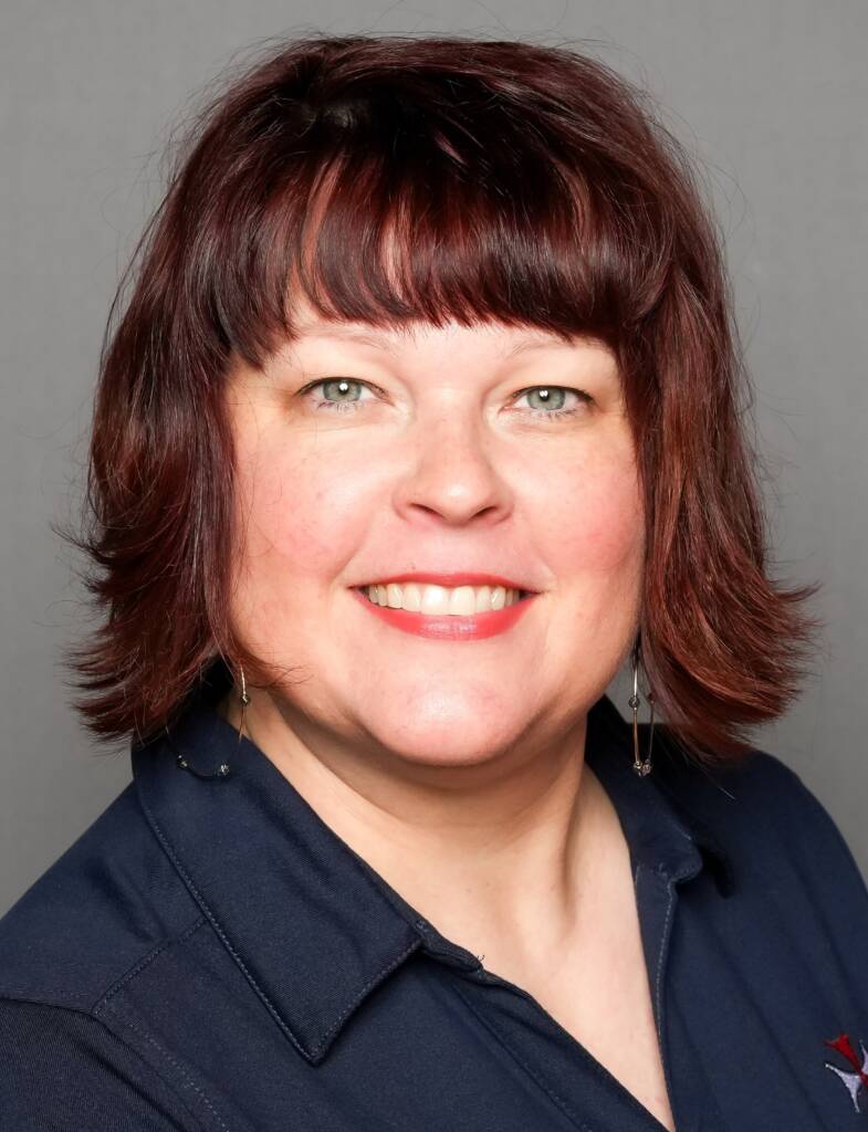 A woman with shoulder-length dark auburn hair and bangs smiles at the camera. She is wearing a navy blue shirt and small dangling earrings. The background is plain and gray.