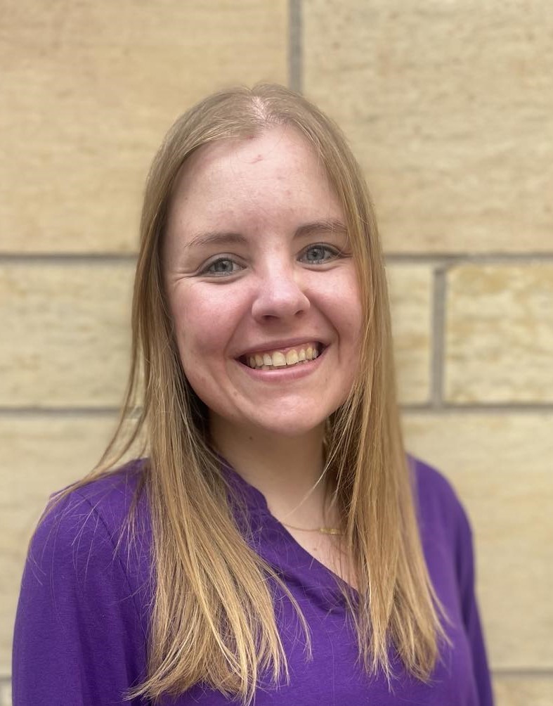 A smiling woman with straight, shoulder-length blond hair wearing a purple top stands in front of a light-colored brick wall.