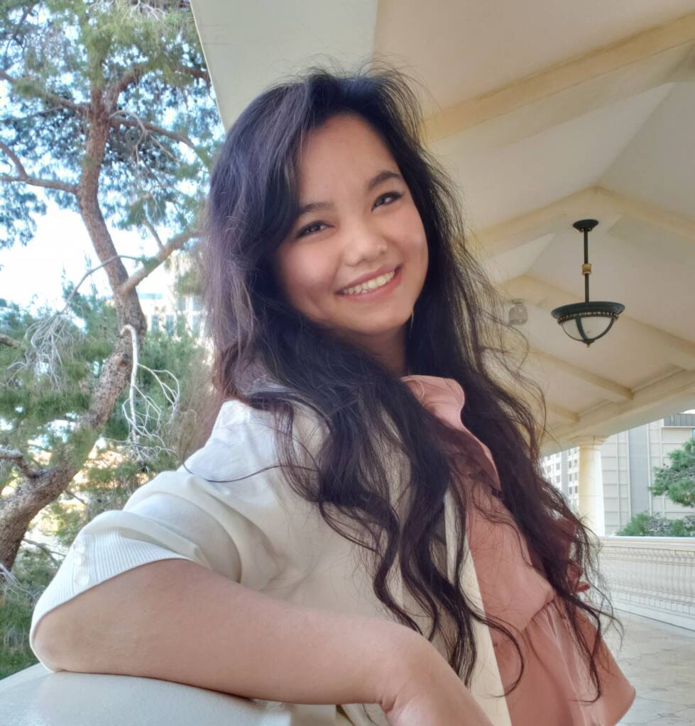 A woman with long wavy hair smiles at the camera, leaning on a white railing. She is wearing a light-colored jacket over a pink blouse. The background features a modern architectural structure and green trees.